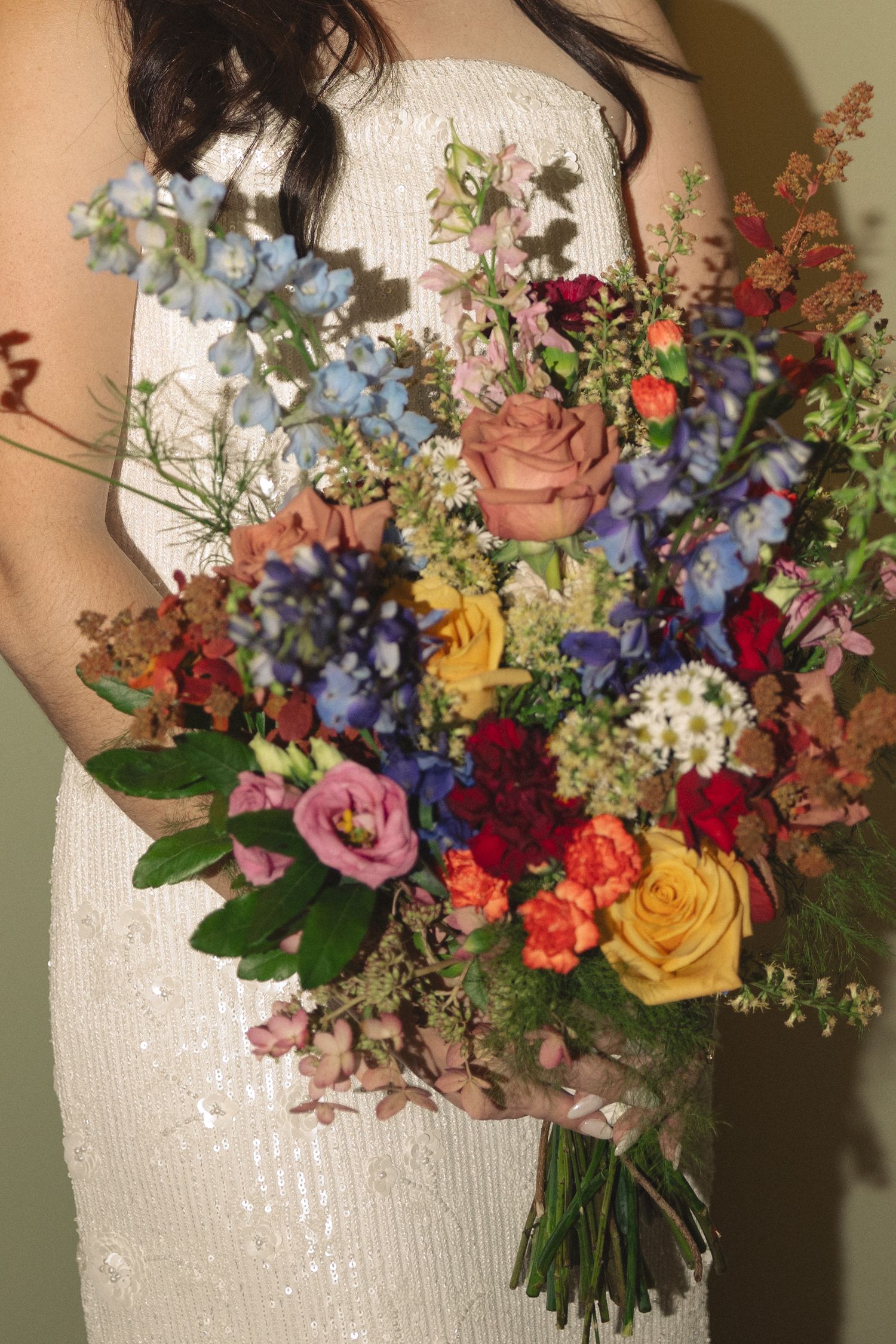 Bride in a white beaded gown holds a colorful bouquet of blue delphinium, roses, ranunculus and wildflowers in orange, pink and red.