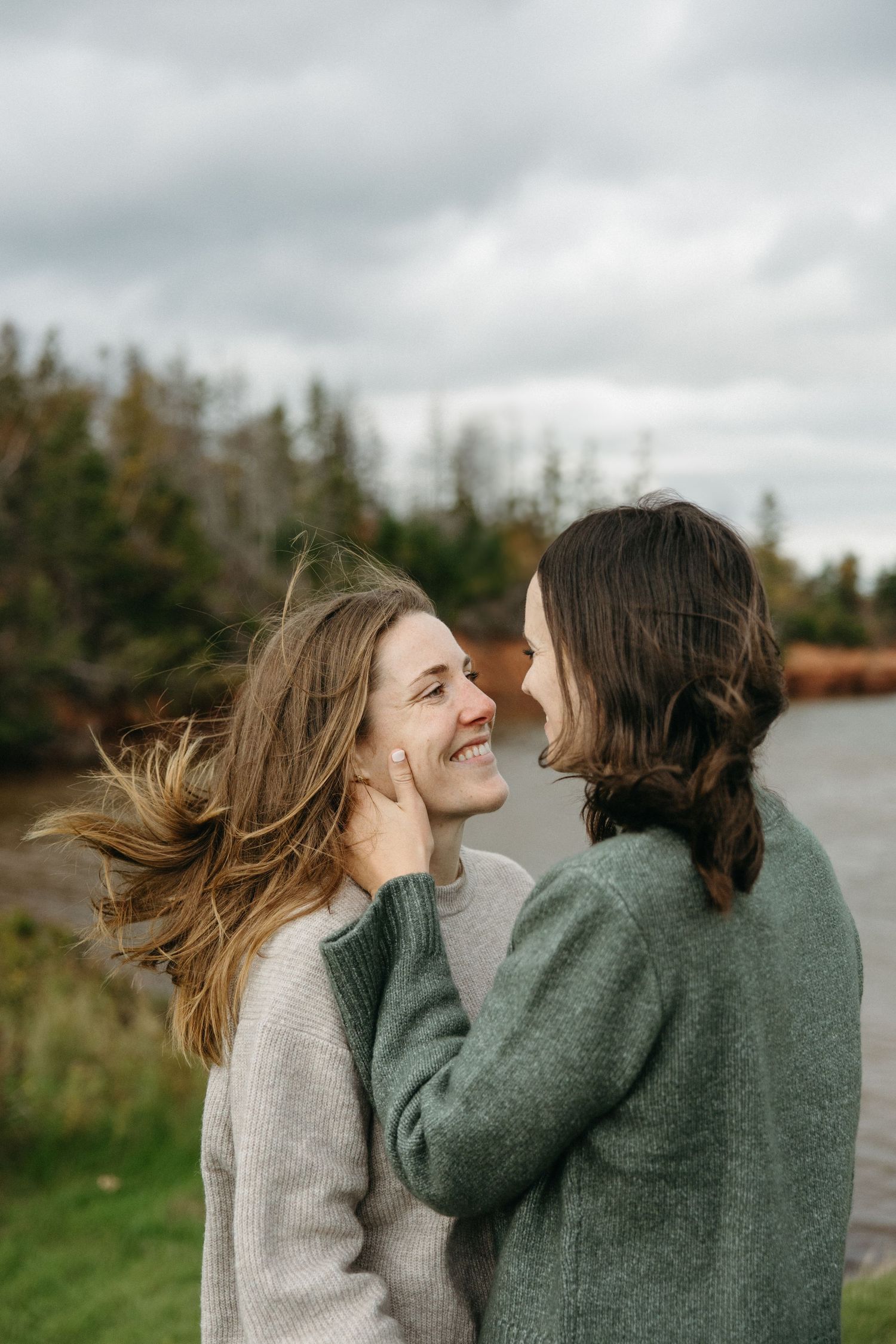 Two women share a joyful moment by a lake, one gently cradling the other's cheek as wind blows her hair.