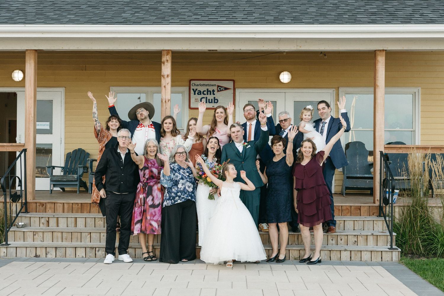 Wedding party with bride in white gown and guests in formal attire cheering and posing on the steps of a yellow wooden lodge.