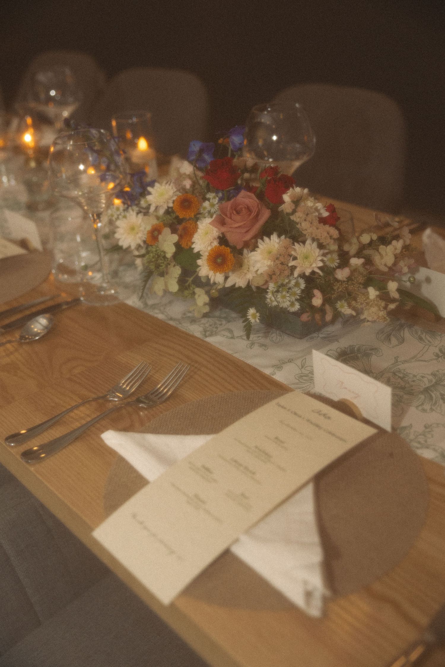 Elegant wedding table setting with a colorful floral centerpiece, candles, wine glasses, silverware, and a printed menu on a wooden table.