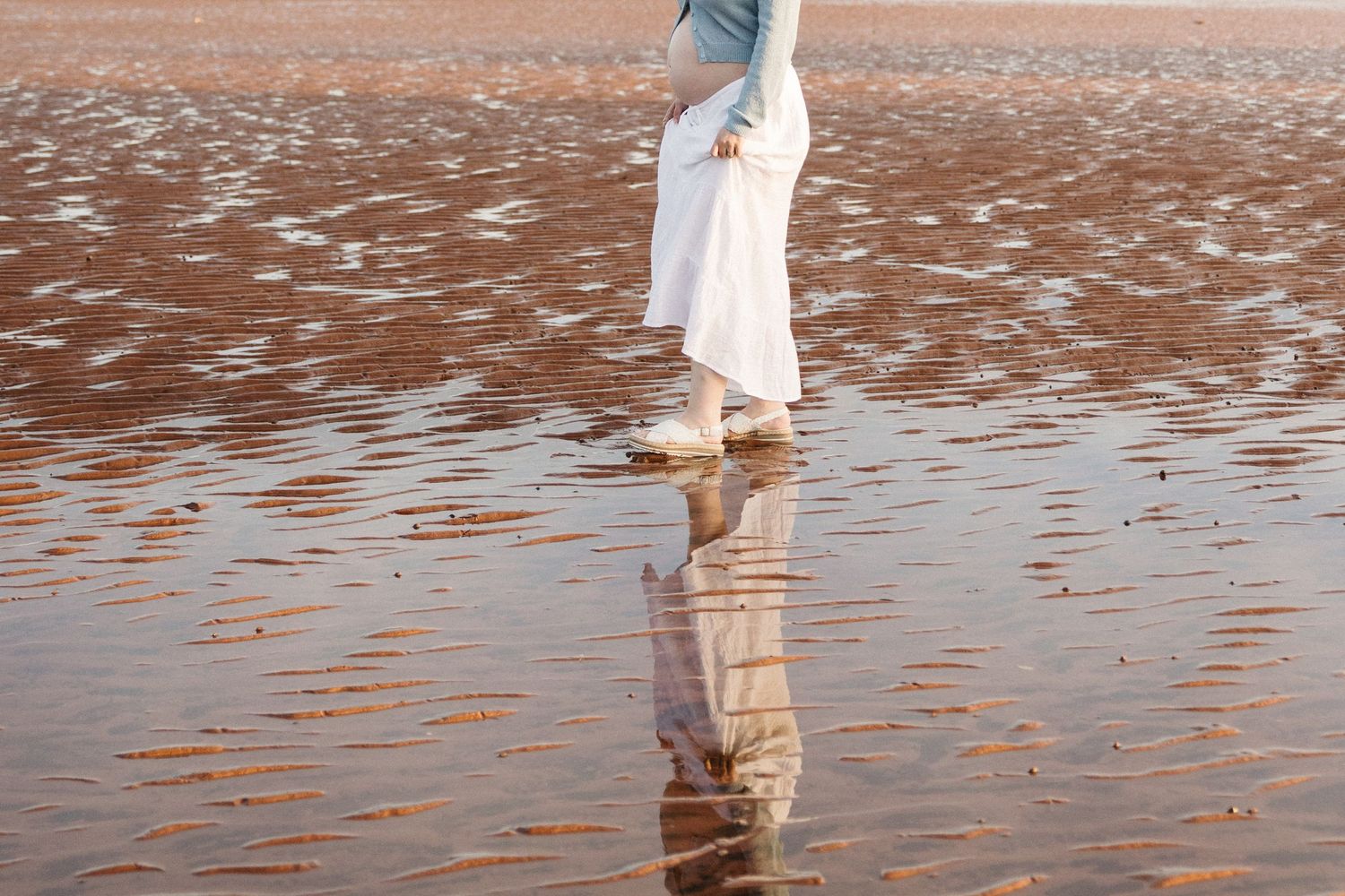 Person in white skirt and denim jacket walking on wet sandy beach with reflection visible in shallow water.