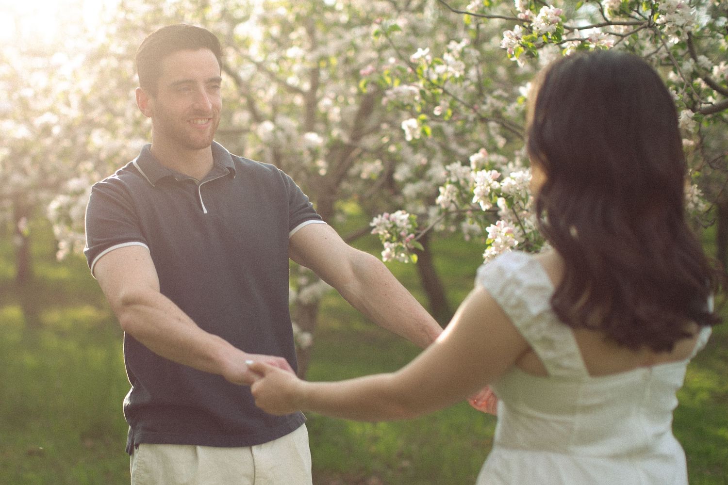 A happy couple holds hands and dances together in a sunny garden with blooming white flowering trees.