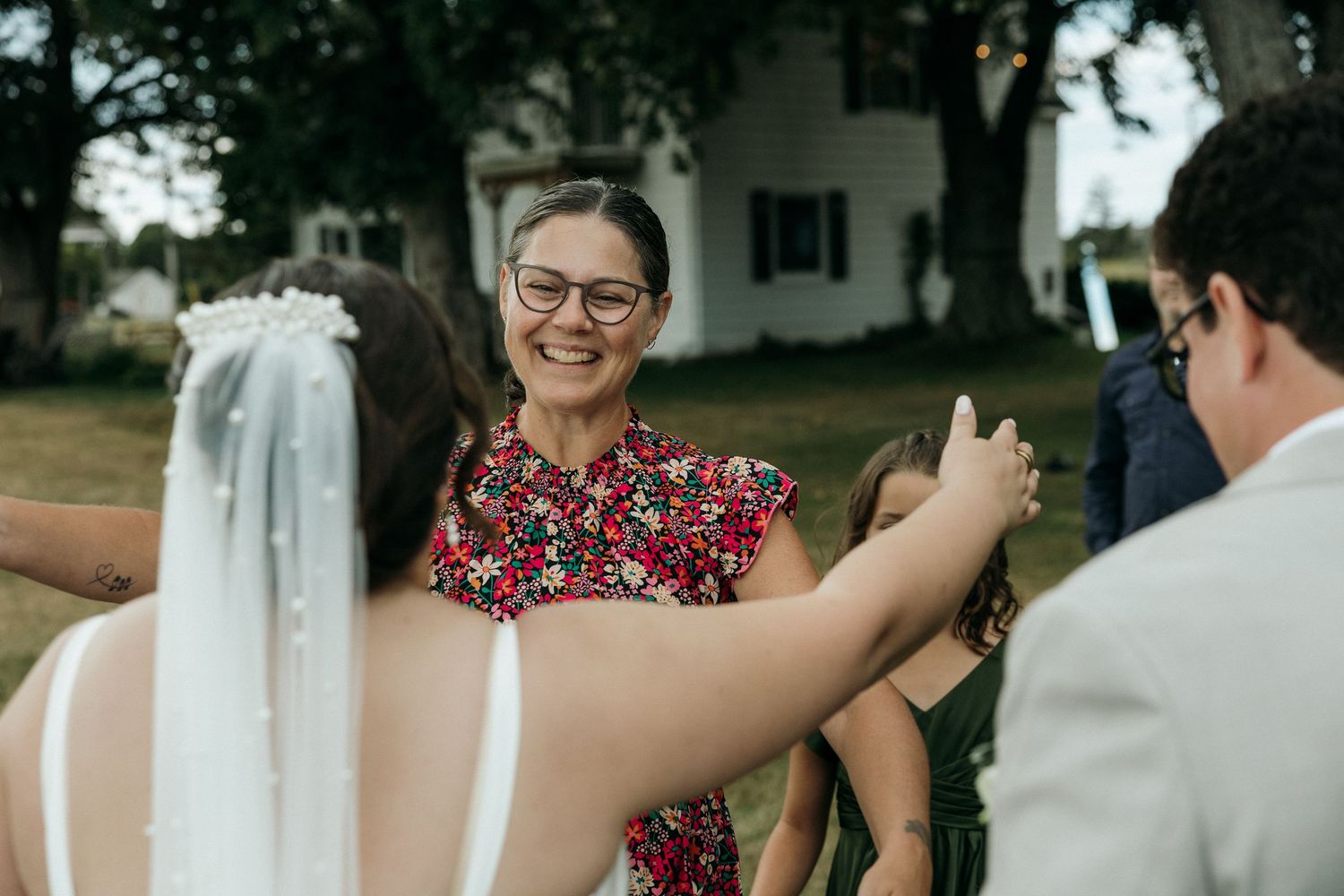 Wedding officiant in floral dress smiles warmly while greeting the bride and groom outdoors.