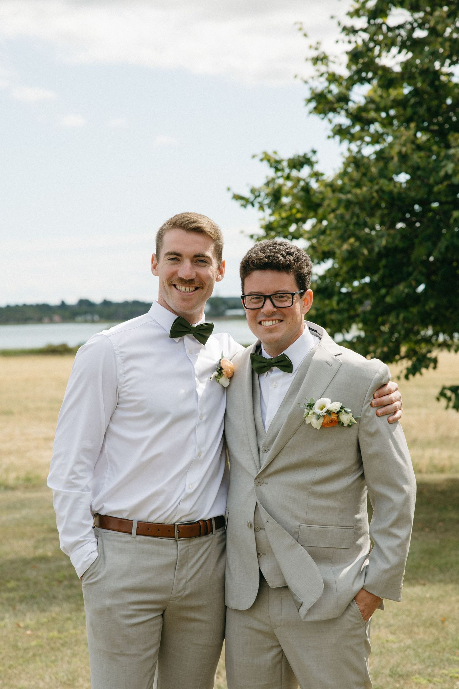 Groom and groomsman smiling outdoors in matching grey suits with green bow ties and boutonnieres.
