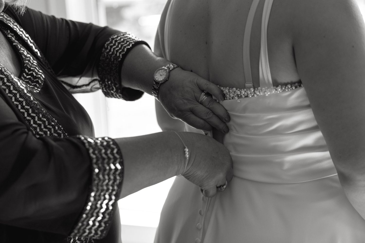Black and white photo of hands fastening the back of a beaded bridal gown before the wedding.