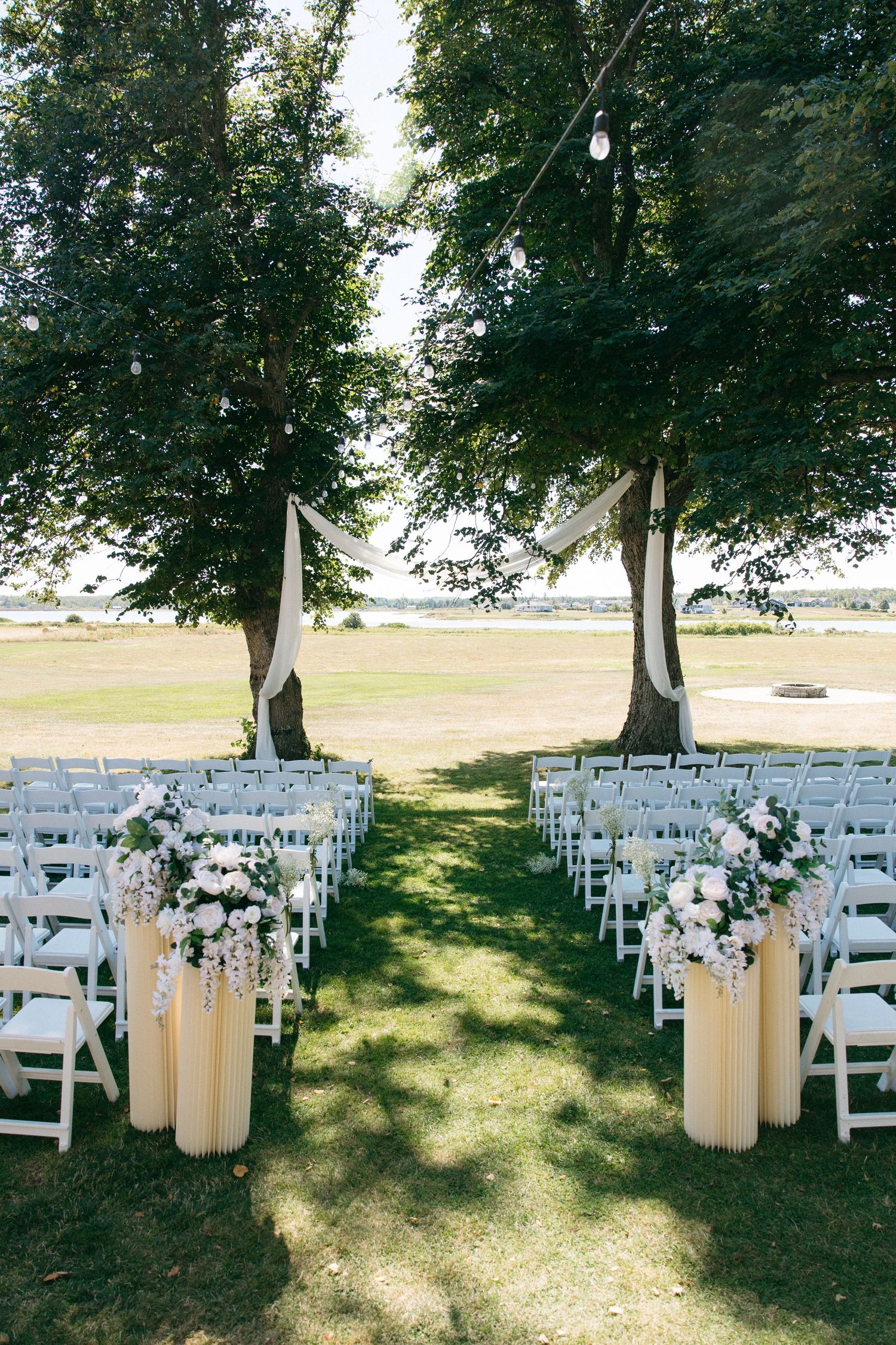 Elegant outdoor wedding ceremony setup with white chairs, floral arrangements, and fabric draped between trees overlooking a waterfront.