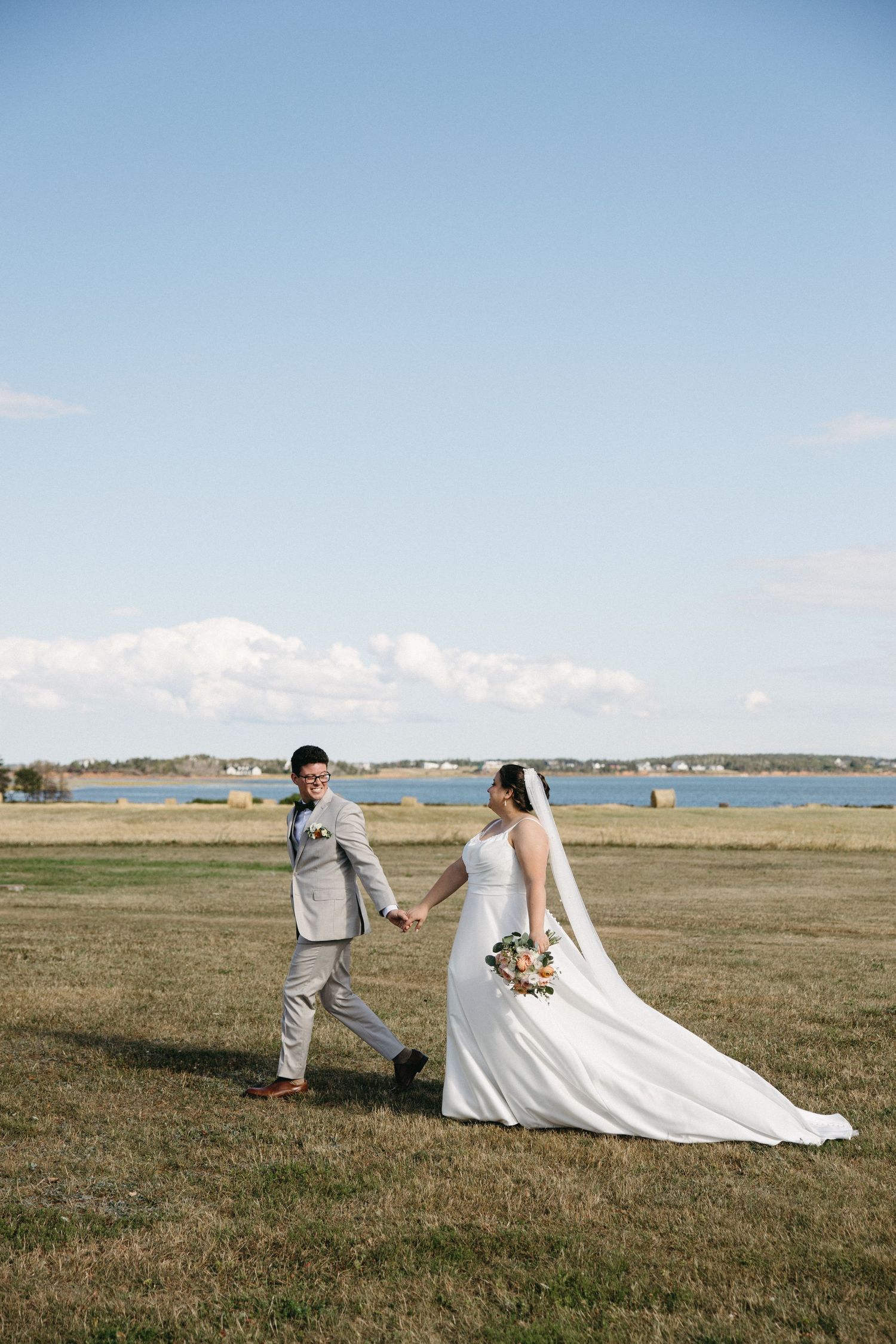 Bride and groom holding hands walking across a scenic waterfront field on their wedding day.
