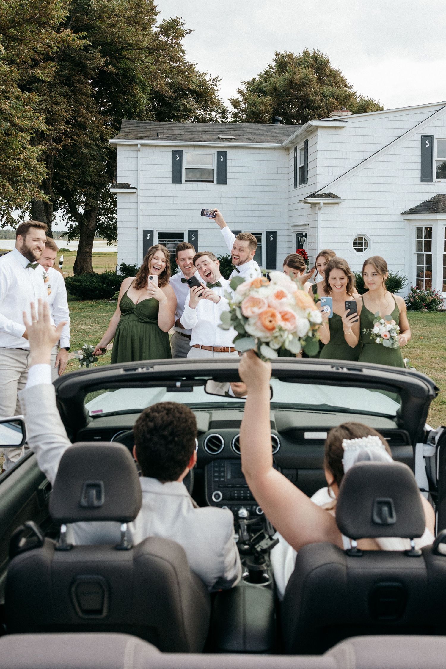 Bride and groom departing in convertible as wedding party waves goodbye outside white farmhouse.