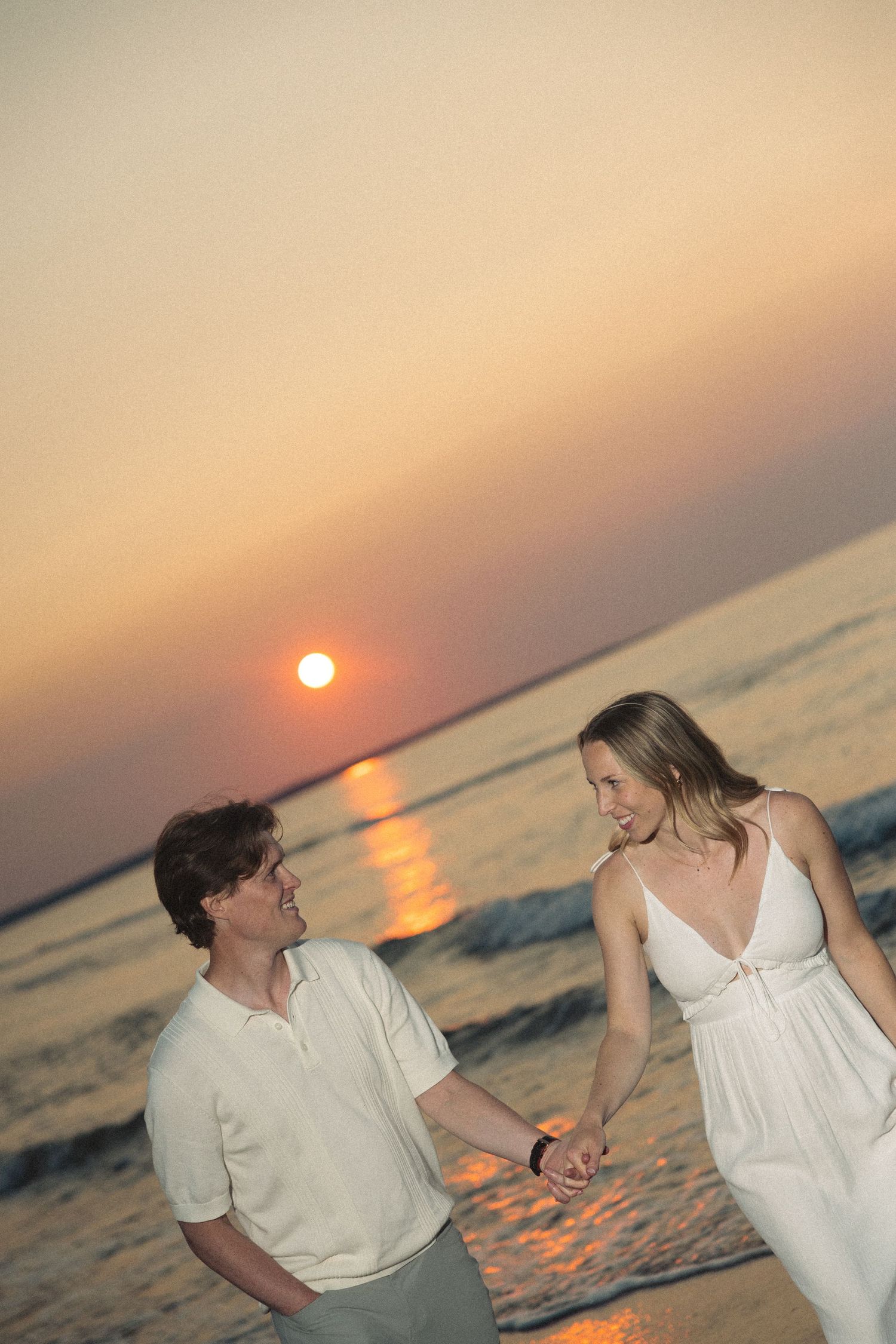 Happy couple holding hands walking on beach at sunset with orange sun reflecting on ocean water.