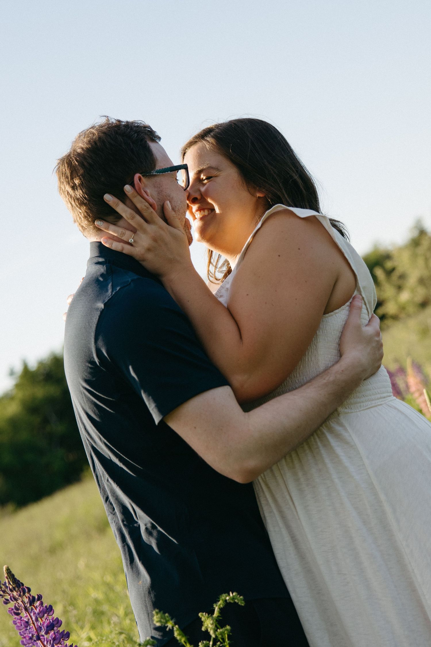 A smiling couple shares a romantic kiss outdoors surrounded by wildflowers and lush greenery at sunset.