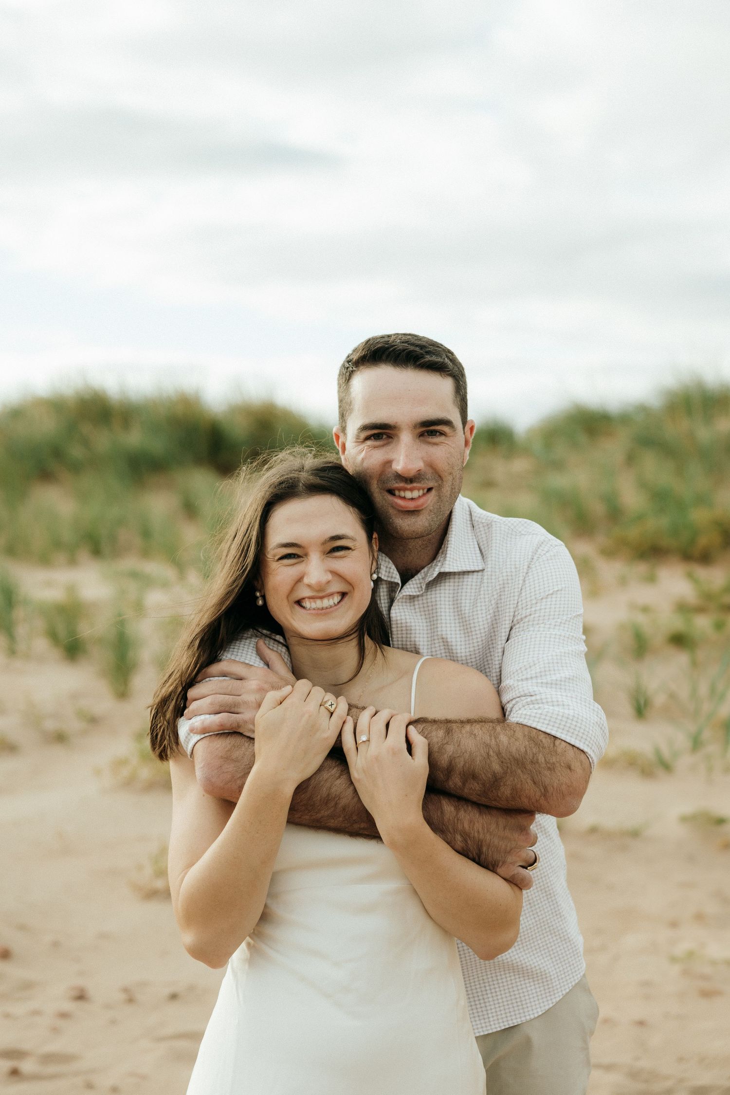 A joyful couple shares a tender moment while standing on a beach with dunes behind them.