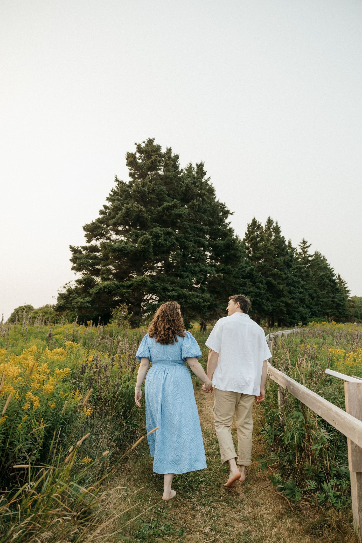 Couple walking hand in hand along a wooden boardwalk through a field of yellow wildflowers.