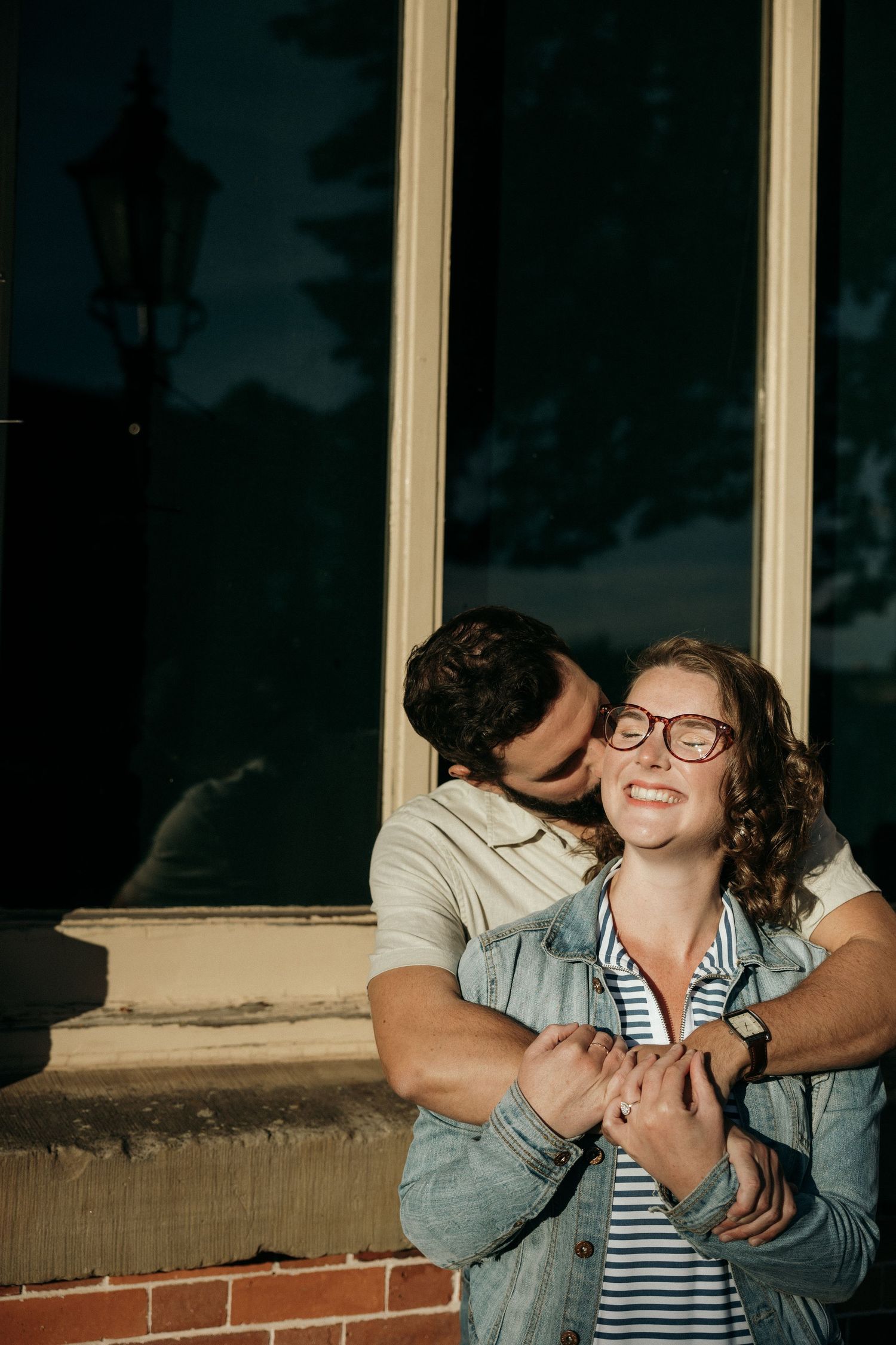 A couple shares a tender embrace near a window with lamp post reflection.