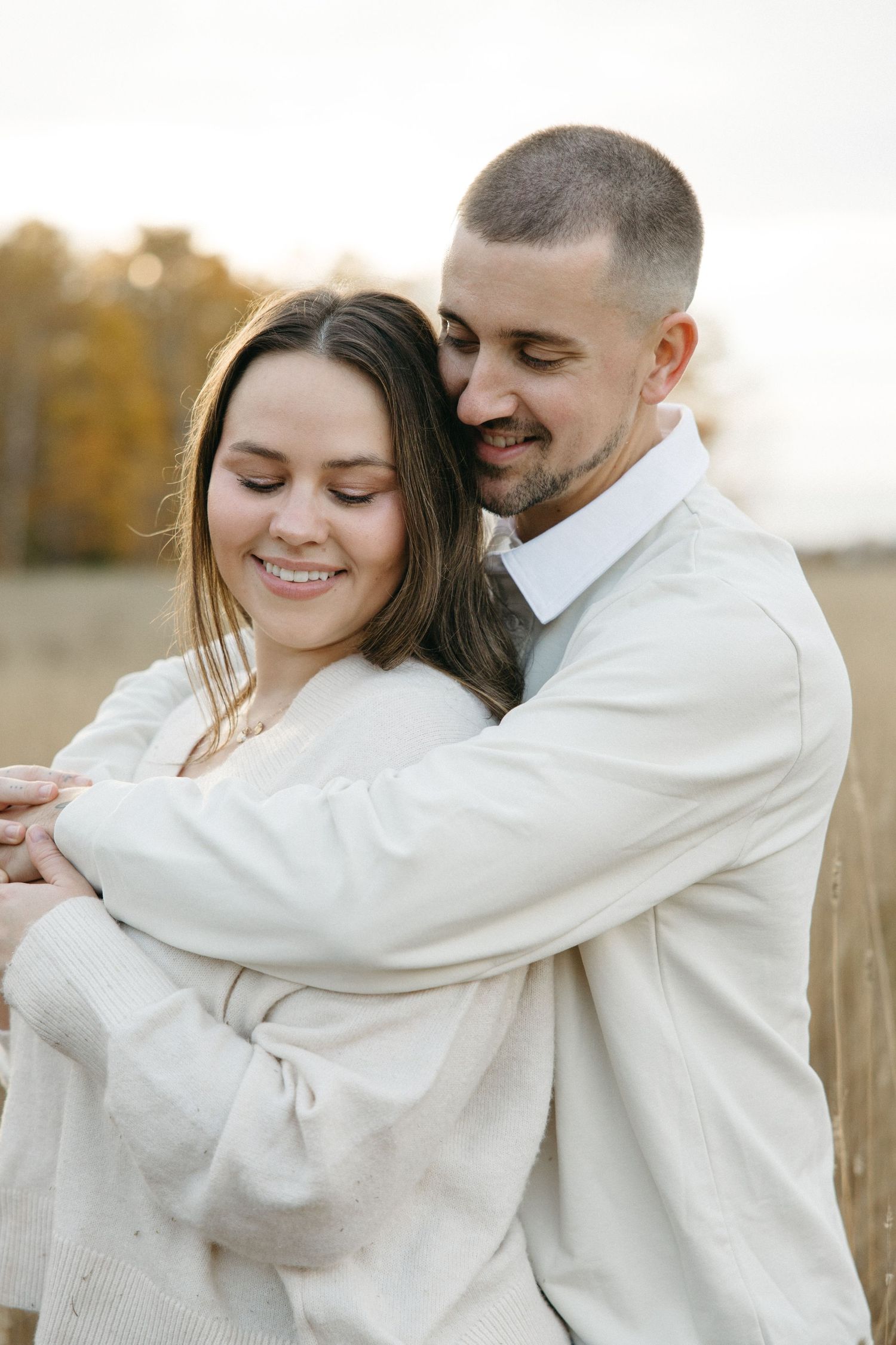 Intimate moment captured as couple embraces in cream sweaters against fall landscape.