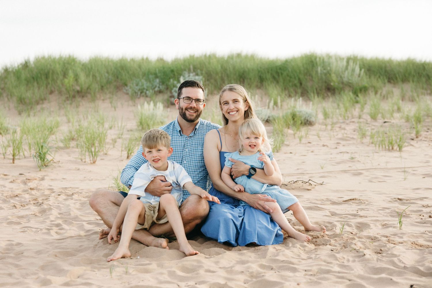 Happy family of four sitting together on sandy beach dunes smiling.