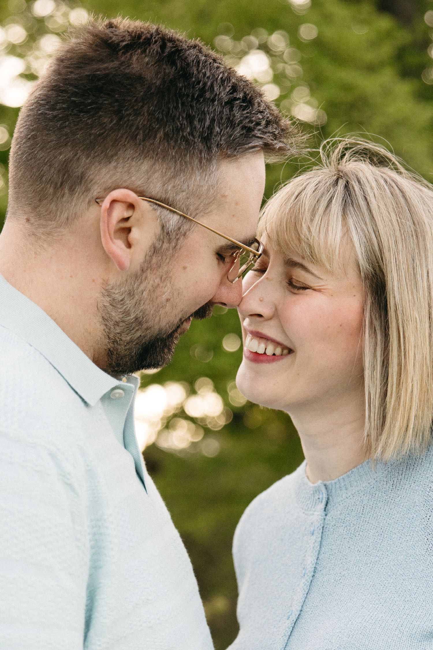 Close-up of smiling couple nearly kissing, both wearing glasses, surrounded by soft green bokeh background.