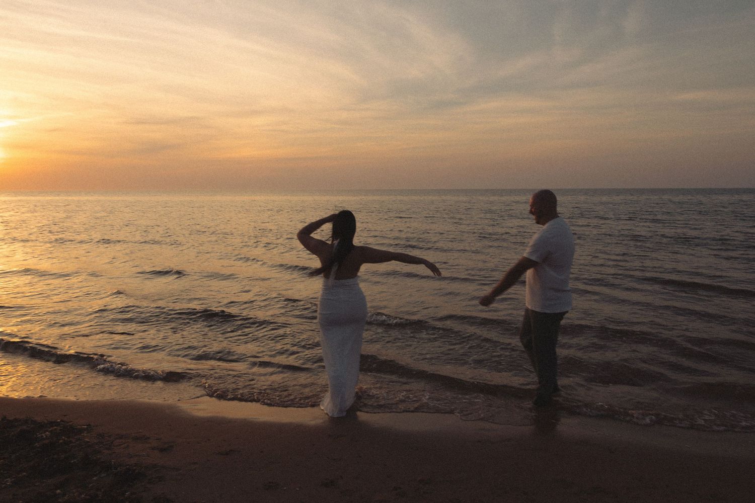 Couple holding hands on a beach shore during a vibrant orange and pink sunset over calm water.