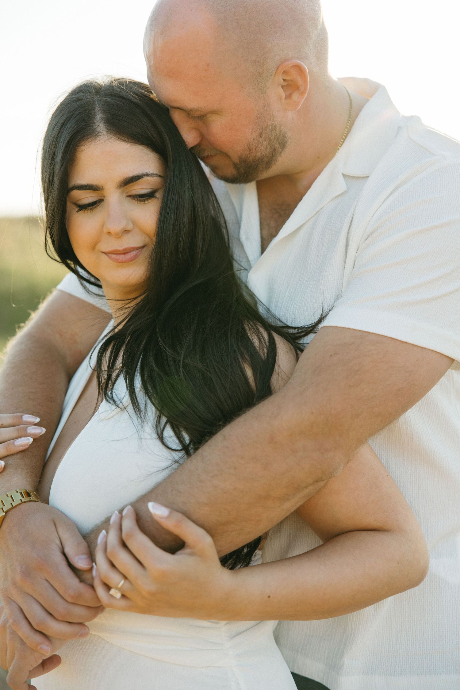 Couple embracing tenderly at golden hour, woman in white dress with engagement ring, man in white shirt hugging her from behind.