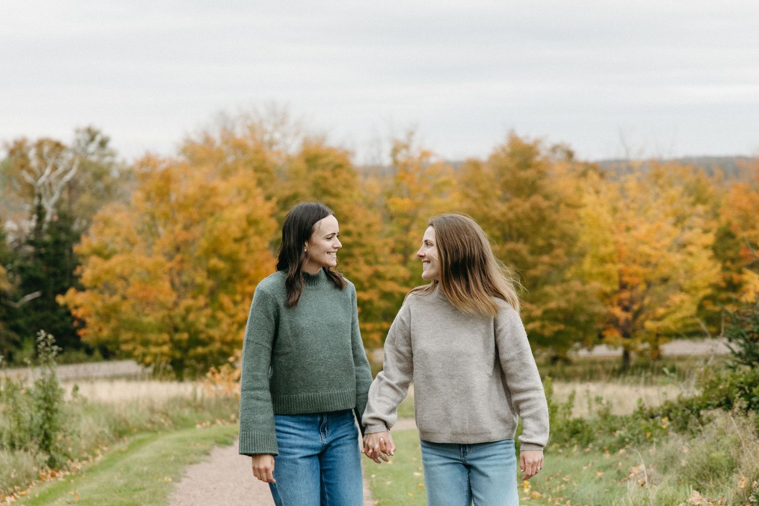 Two women hold hands and smile while walking along a grassy path with vibrant orange and yellow autumn trees in the background.