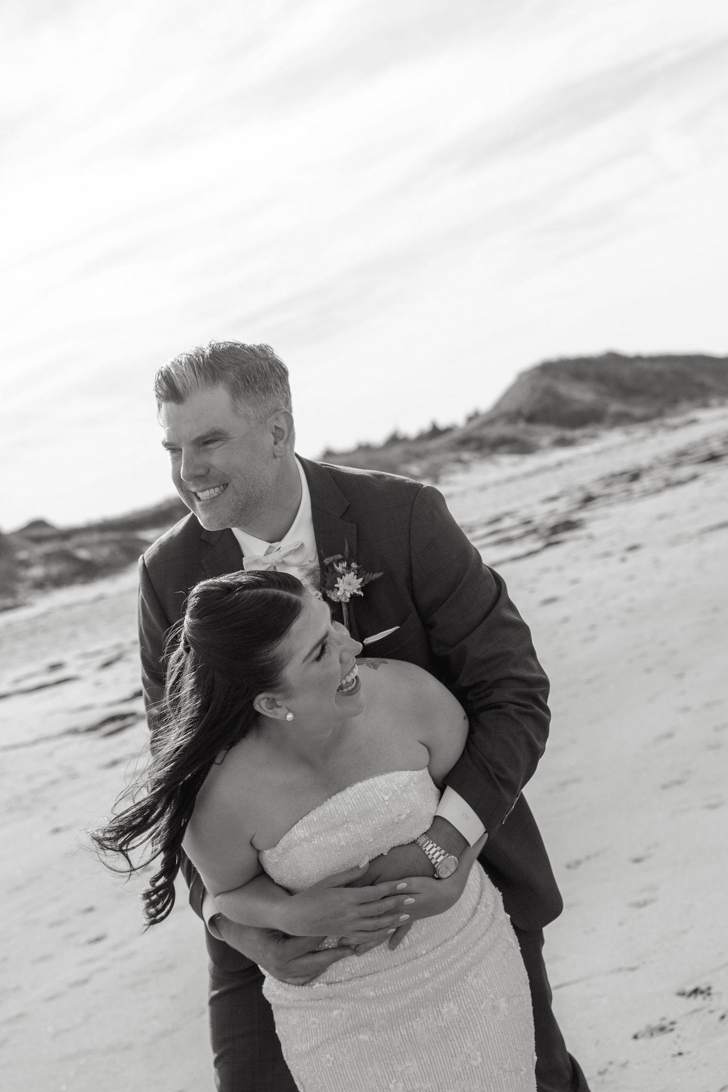 Black and white photo of a laughing groom embracing his bride as the wind blows her hair on a coastal beach.