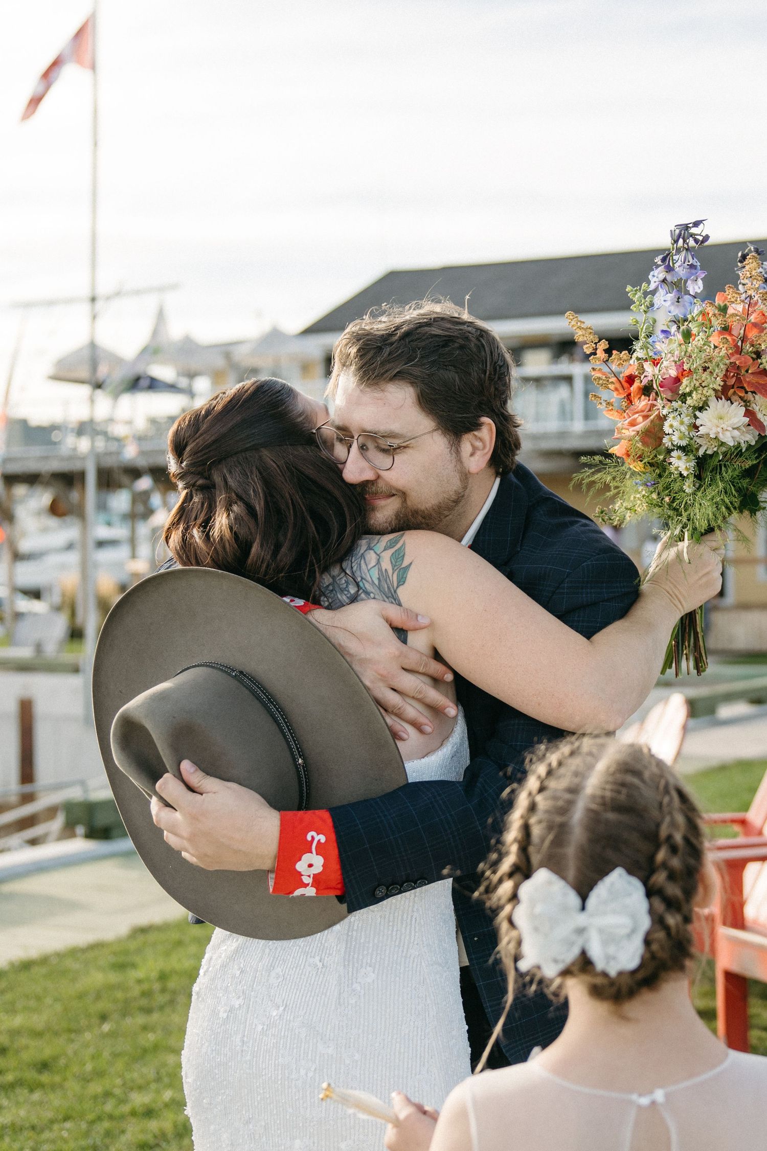 Bride in a lace wedding dress holding a gray hat embraces a bearded groom with glasses as a flower girl in a bow watches nearby.