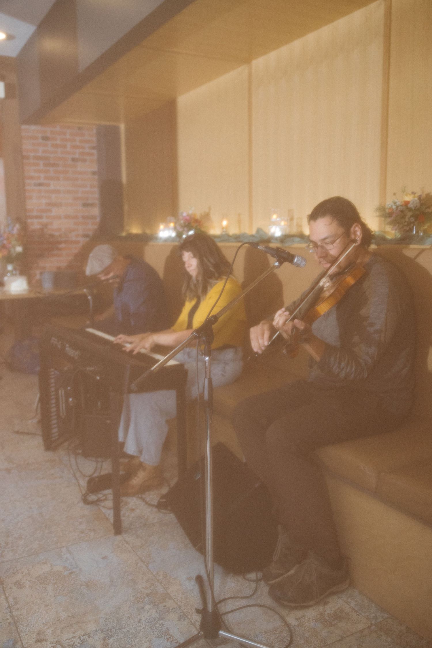 Three musicians perform live in a warmly lit lounge, with a violinist and a keyboardist seated on a bench beside candles and flowers.