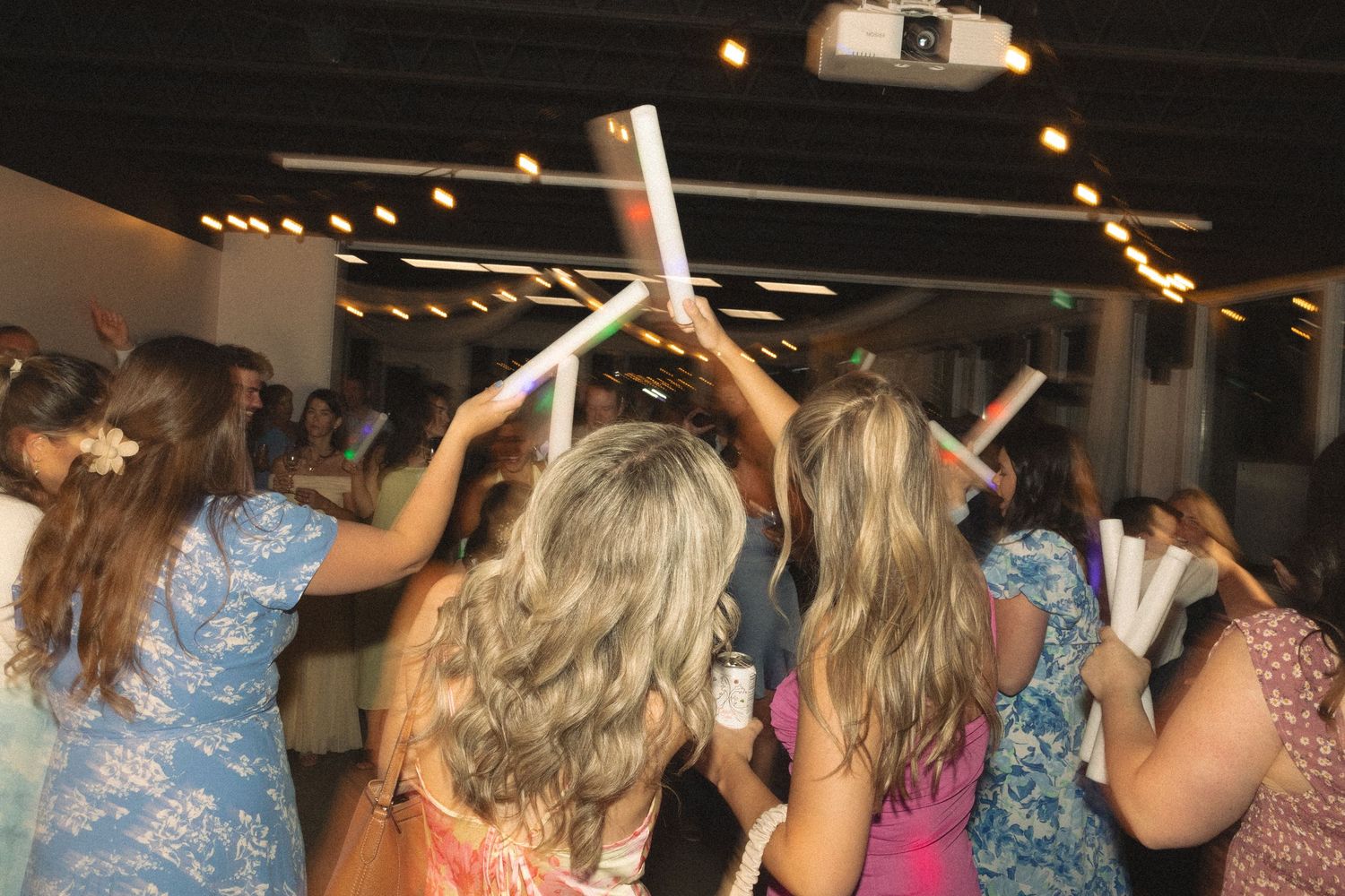Wedding guests dance with arms raised under string lights at an indoor reception.