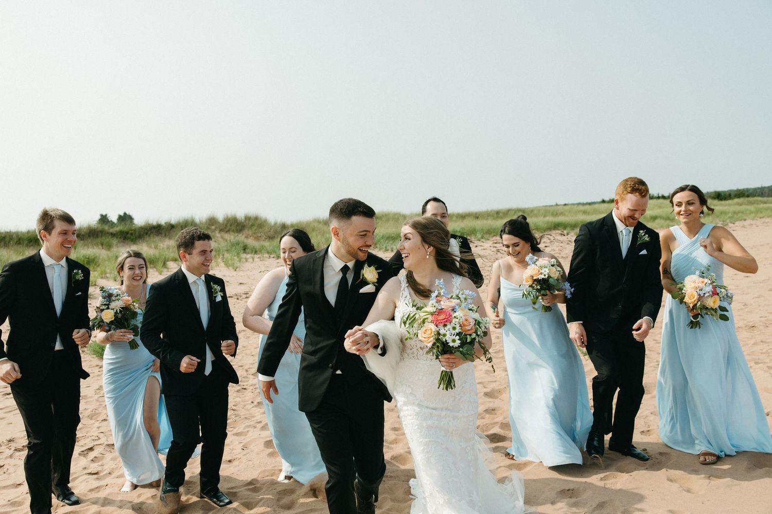 Wedding party laughing and walking together on beach with bridesmaids in light blue dresses.