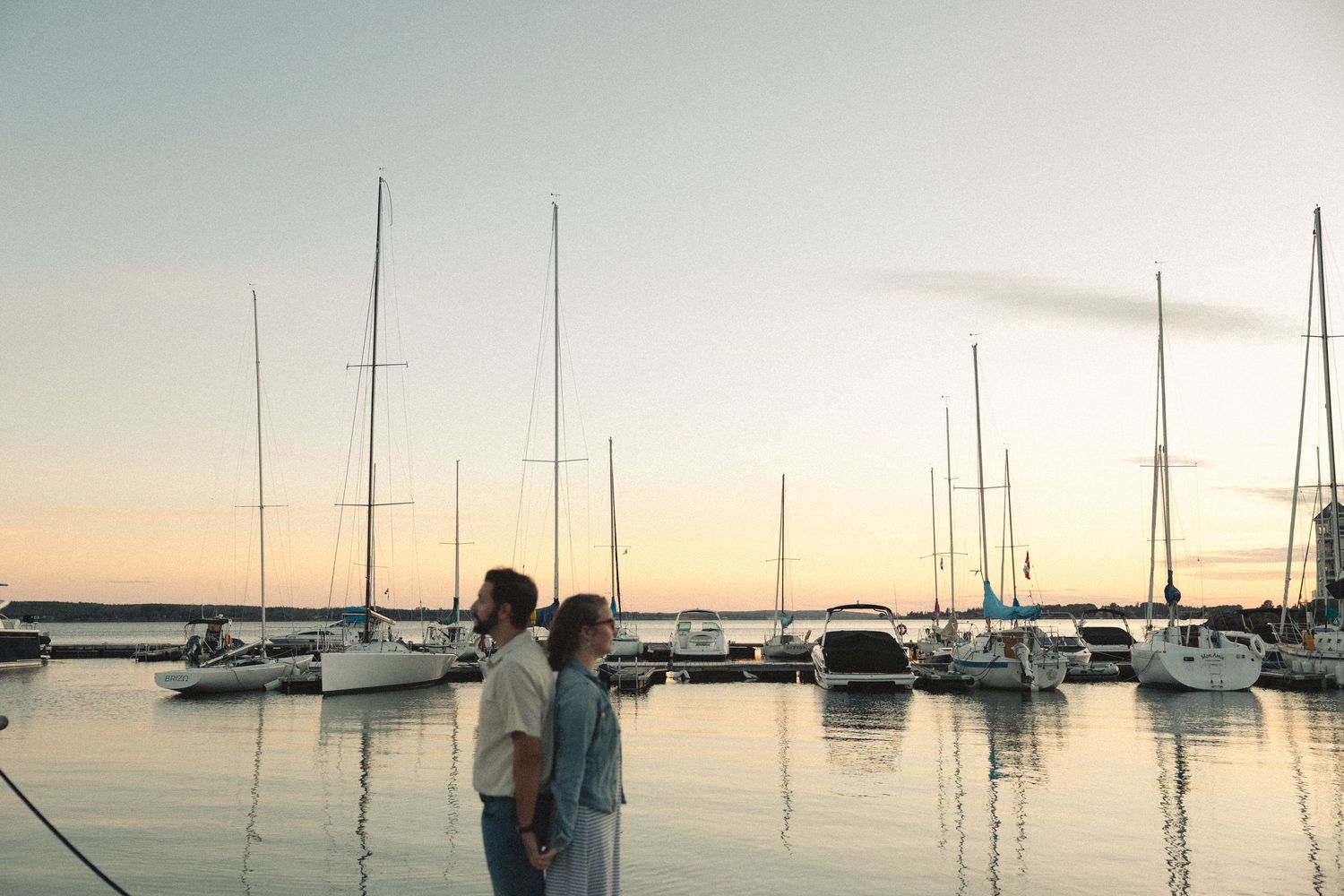 A couple stands together at sunset in front of a marina filled with sailboats.