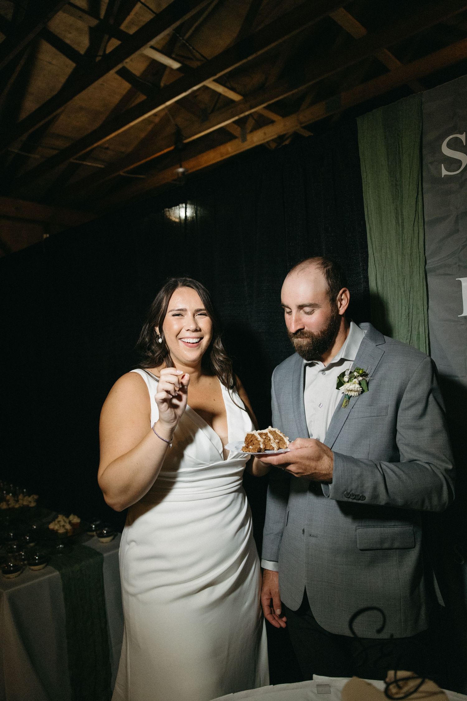 A couple shares a playful moment feeding each other cake at a wedding reception in a rustic barn venue.
