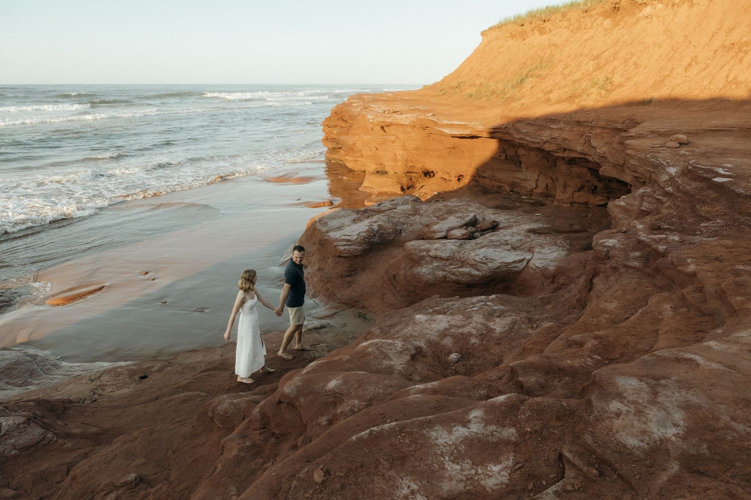 Two people explore rocky coastal cliffs overlooking the ocean at golden hour.