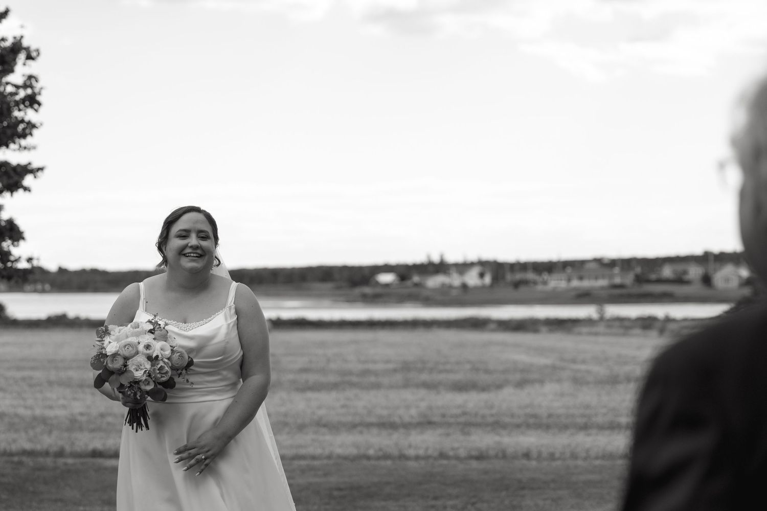 Smiling bride in white gown holding bouquet in black and white outdoor wedding photo by waterfront.