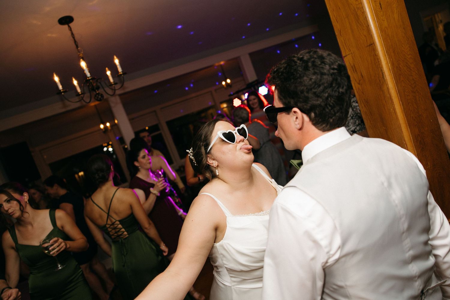 Bride in white dress wearing novelty sunglasses dances with a guest during lively wedding reception celebration.