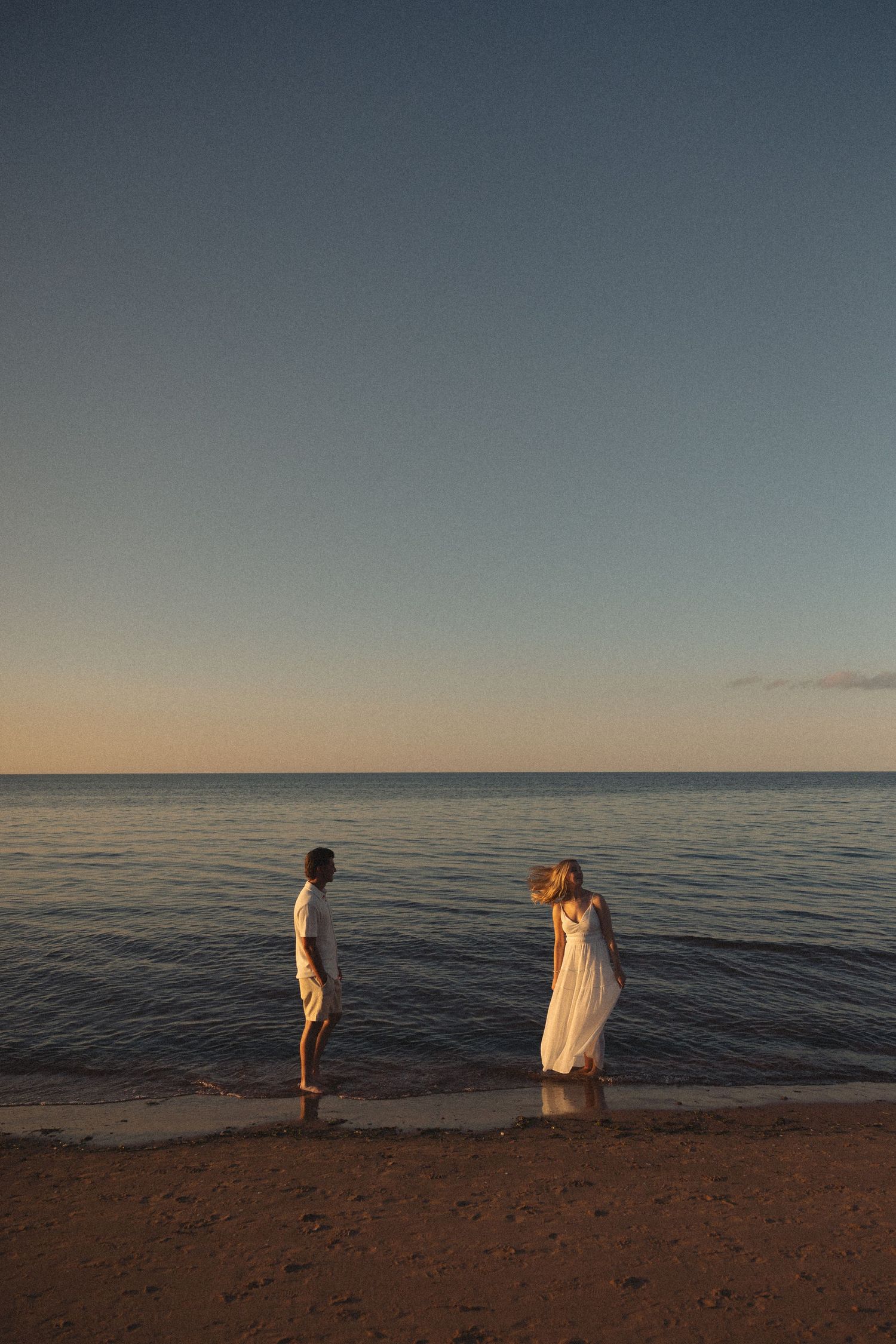 Couple in white outfits standing at the water's edge on a sandy beach during golden hour sunset.