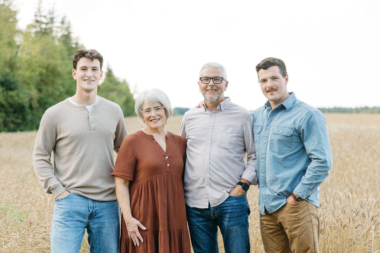Family of four smiling together in a golden wheat field with trees in the background.