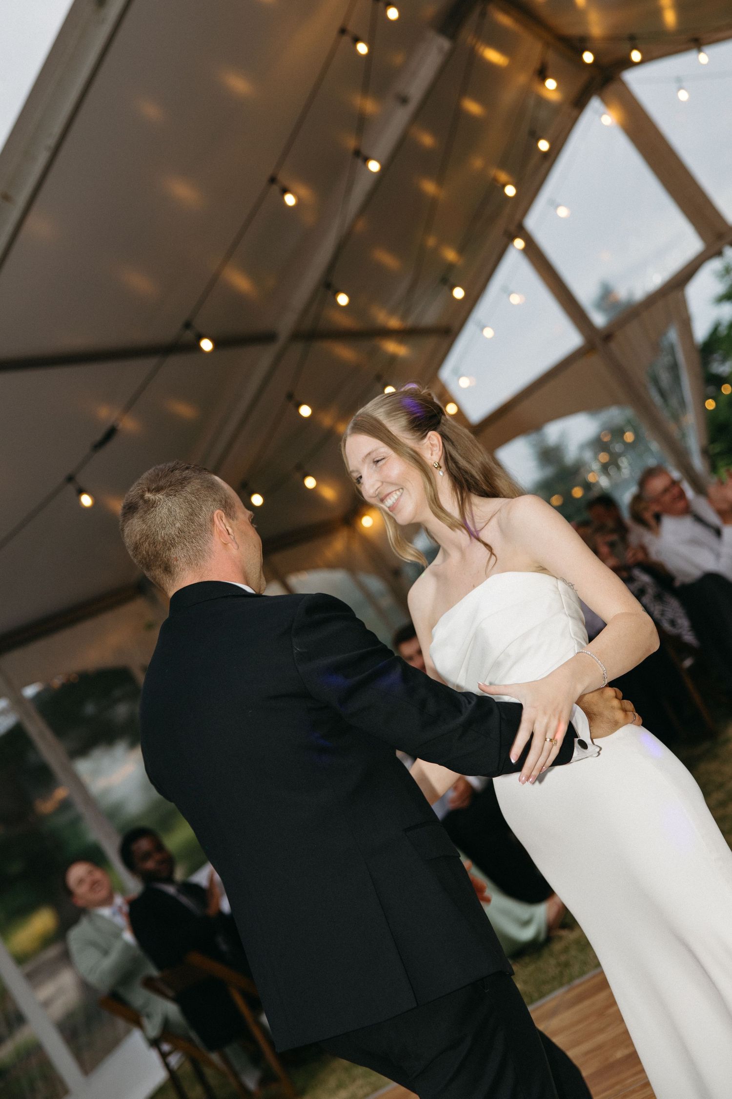 Bride in strapless white gown laughs joyfully while sharing first dance under string lights in a wedding tent.