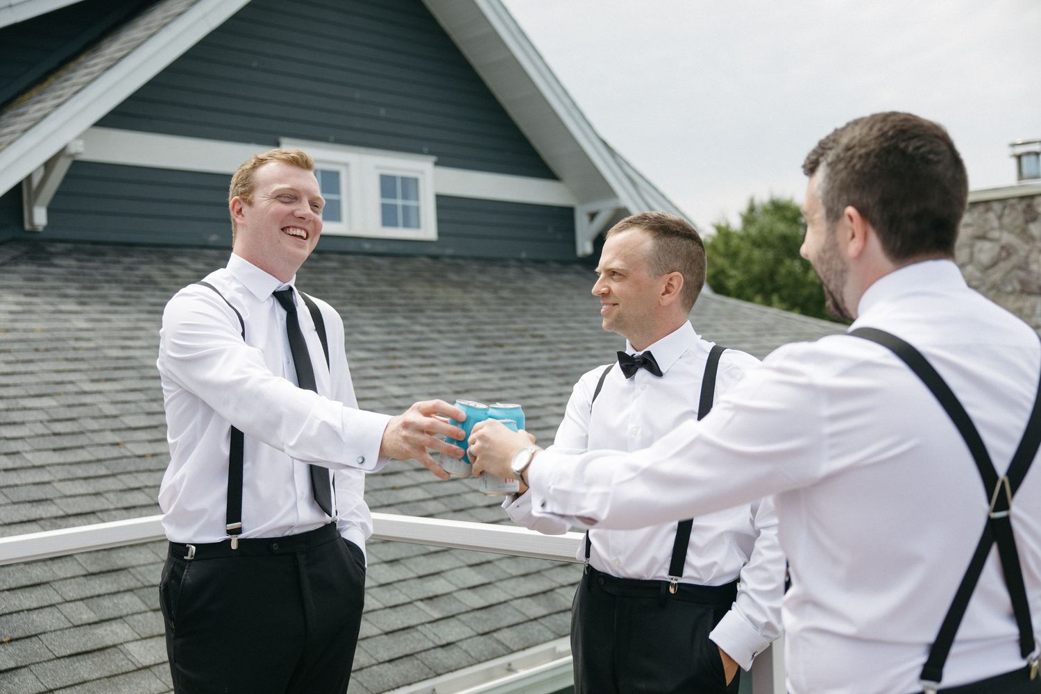 Three groomsmen in white shirts and suspenders share a toast outdoors before a wedding ceremony.