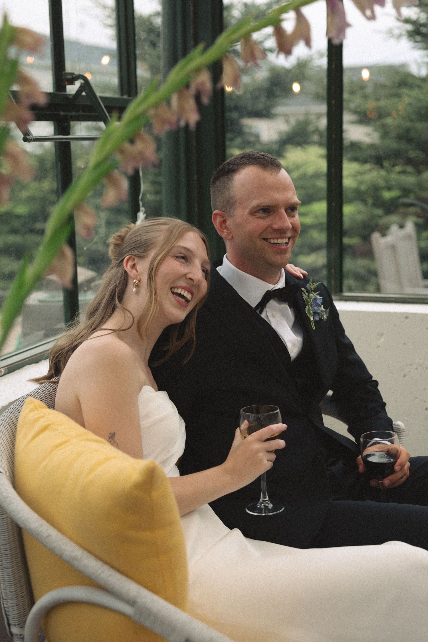 Laughing bride and groom in formal attire relaxing on a couch with wine glasses in a greenhouse setting.