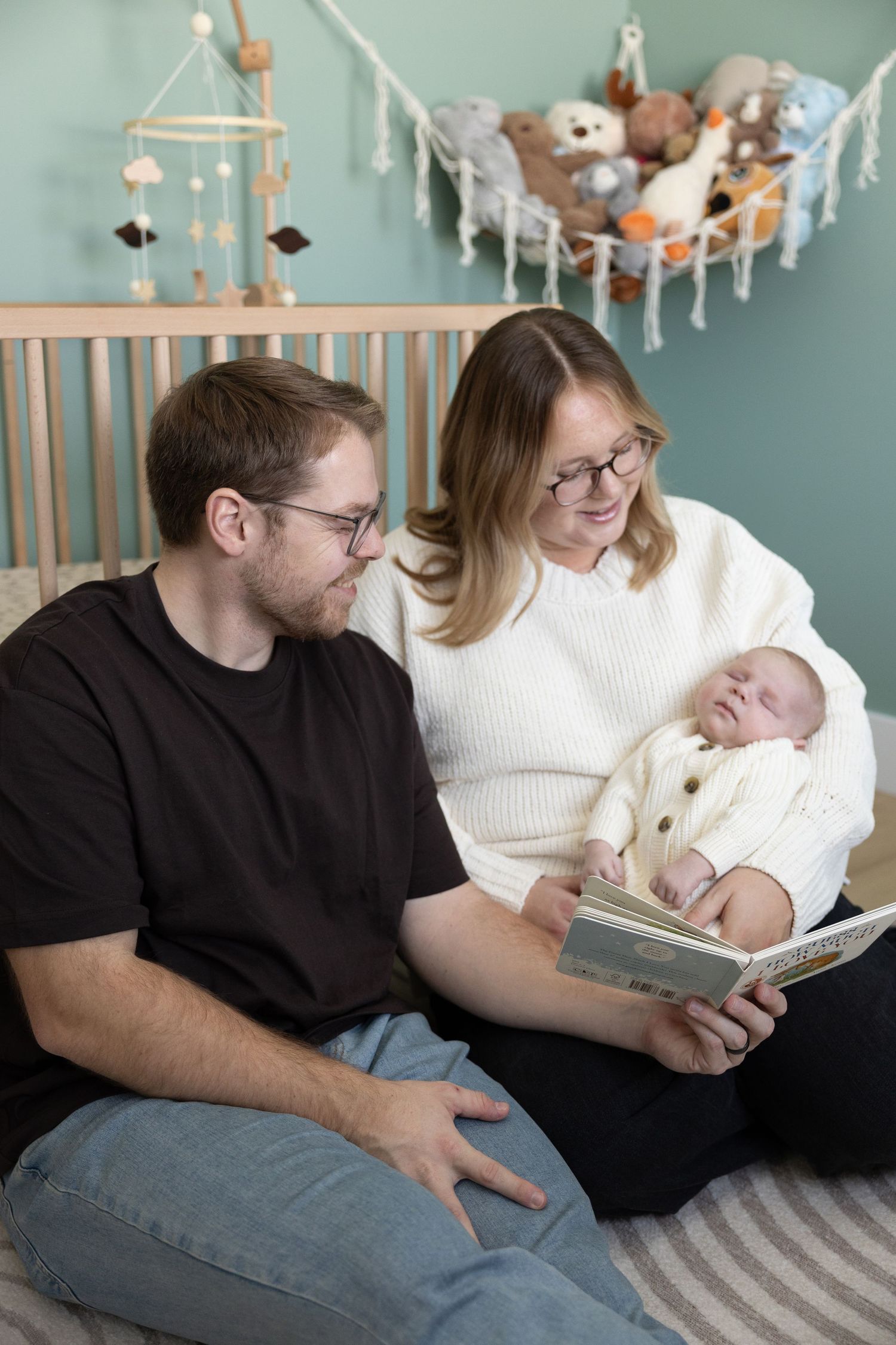 Family relaxing together in nursery with mobile and stuffed animals visible in background.