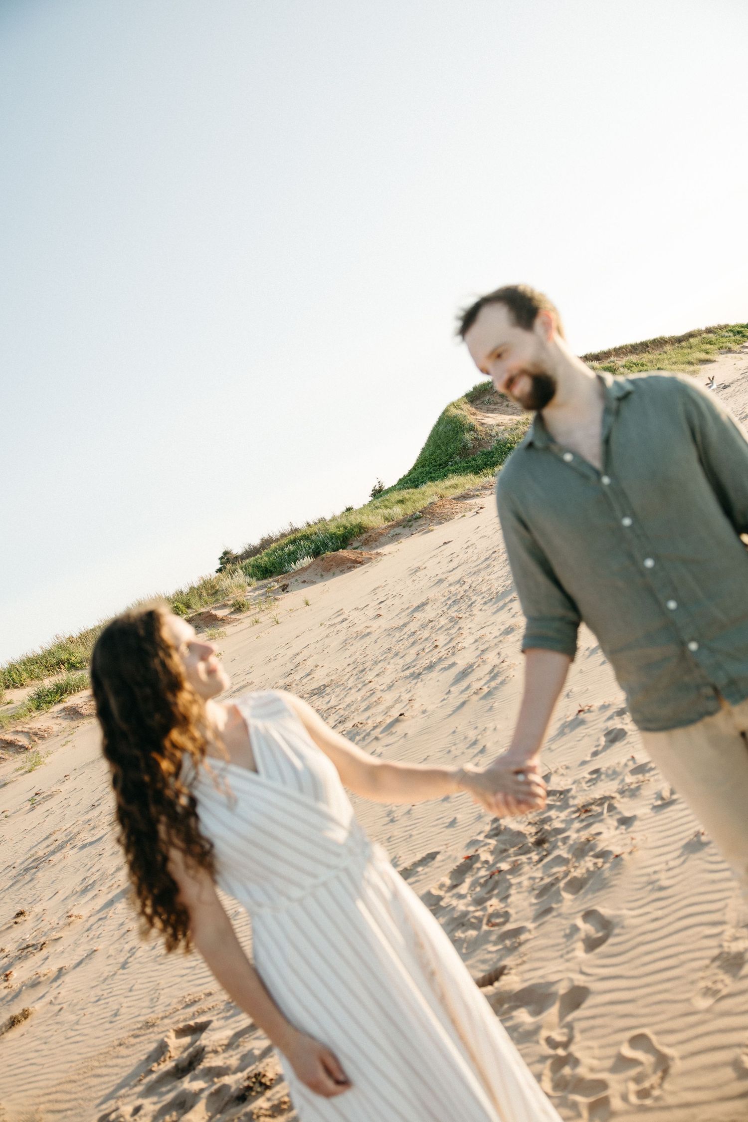 Couple holding hands on sandy beach, man smiling at woman in white striped dress under blue sky.