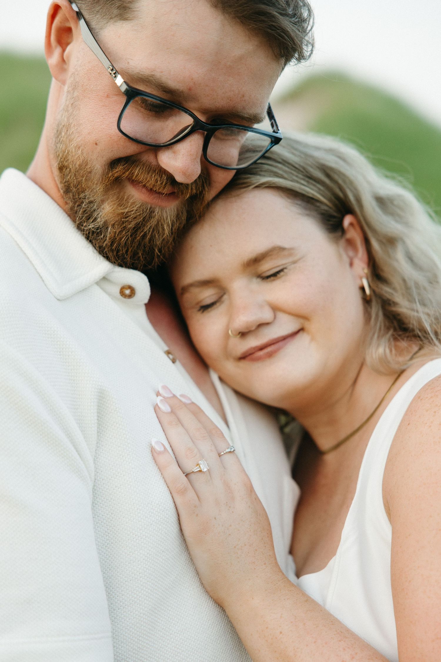 Engaged couple in white outfits share a tender moment, woman displaying diamond ring while resting head on mans chest.