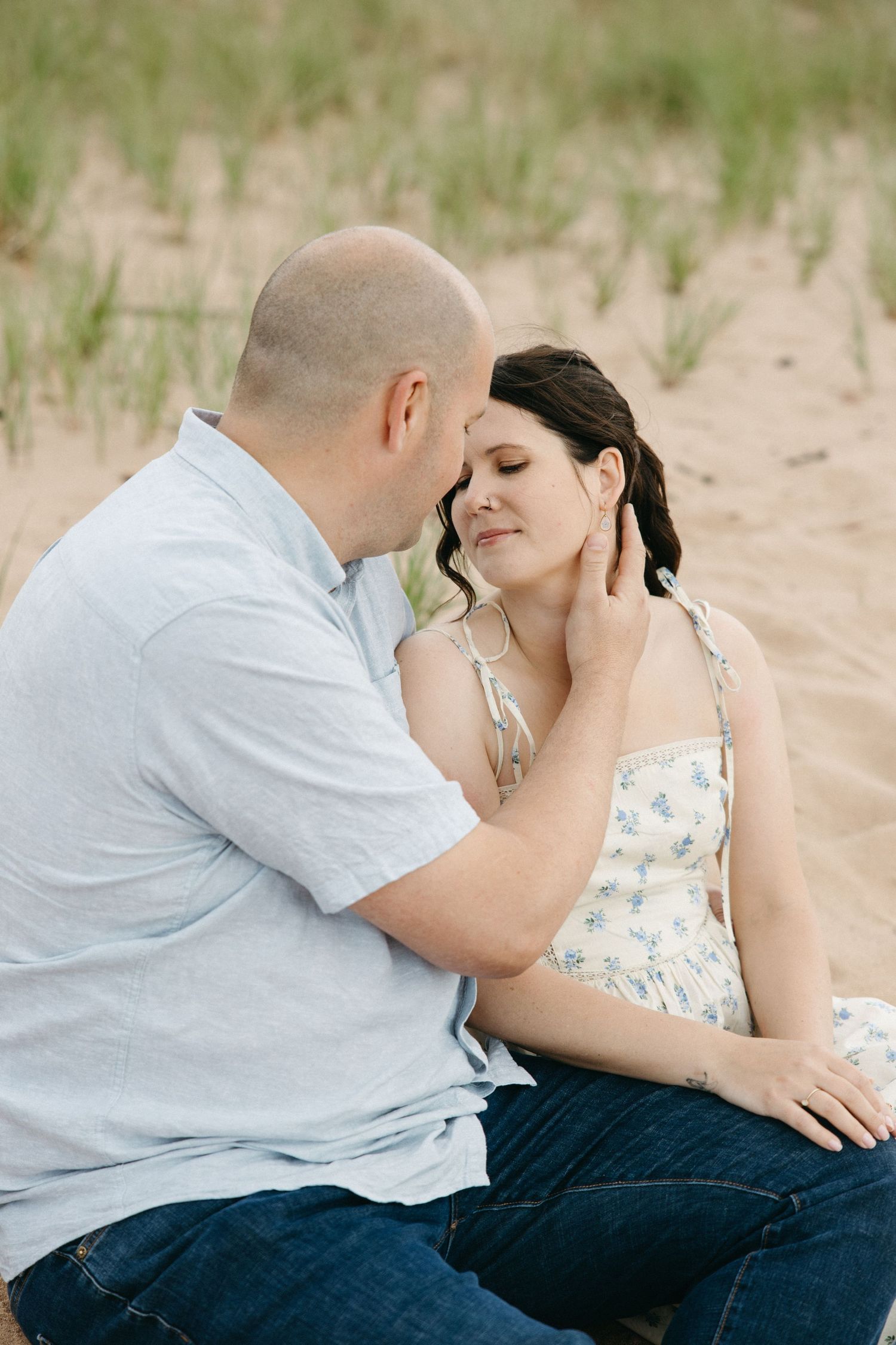A couple sits intimately on sandy beach dunes as the man tenderly touches her face.