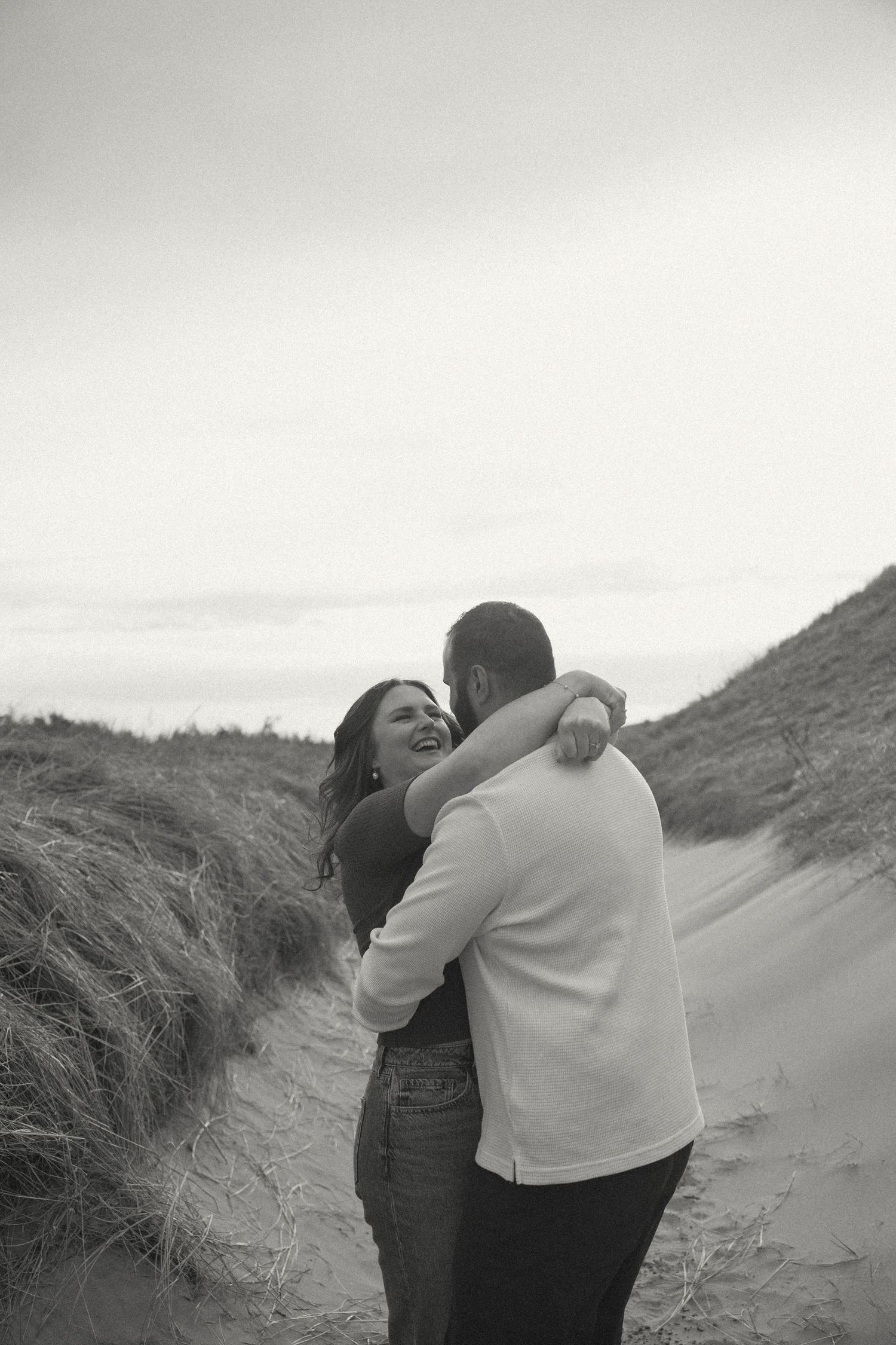 Two people embrace playfully among coastal sand dunes on a cloudy day.