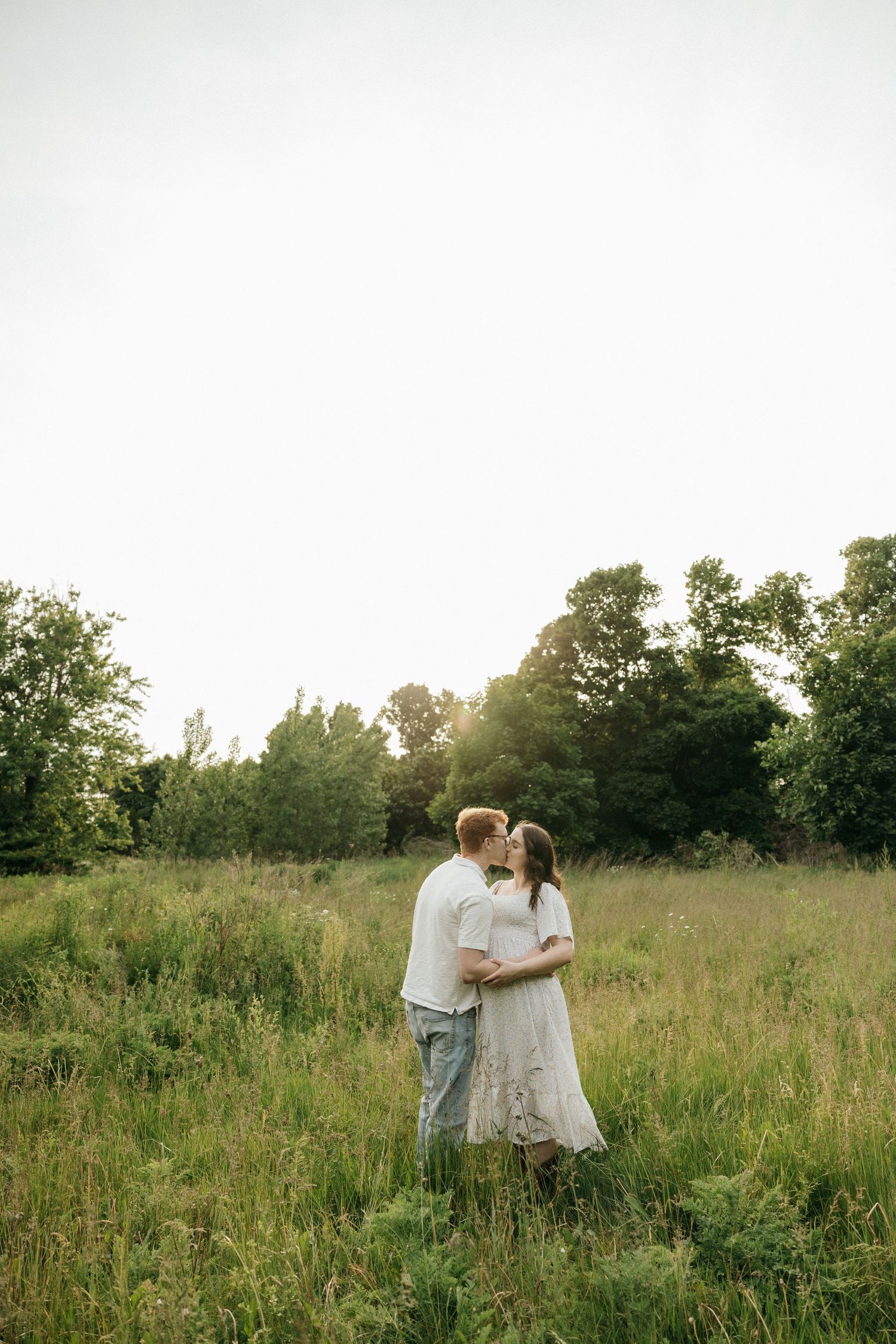 A couple shares a tender moment in a sunlit meadow with tall grass and trees in the background.