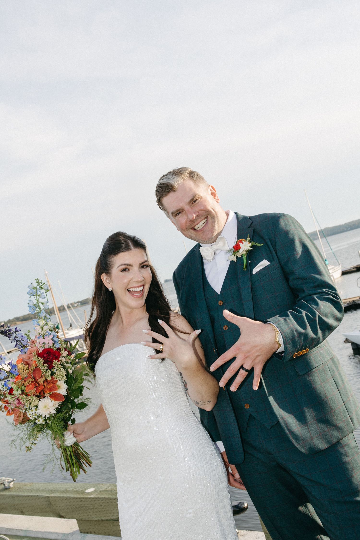 Smiling bride in a white strapless gown and groom in a green suit show their wedding rings by the water, holding a colorful bouquet.