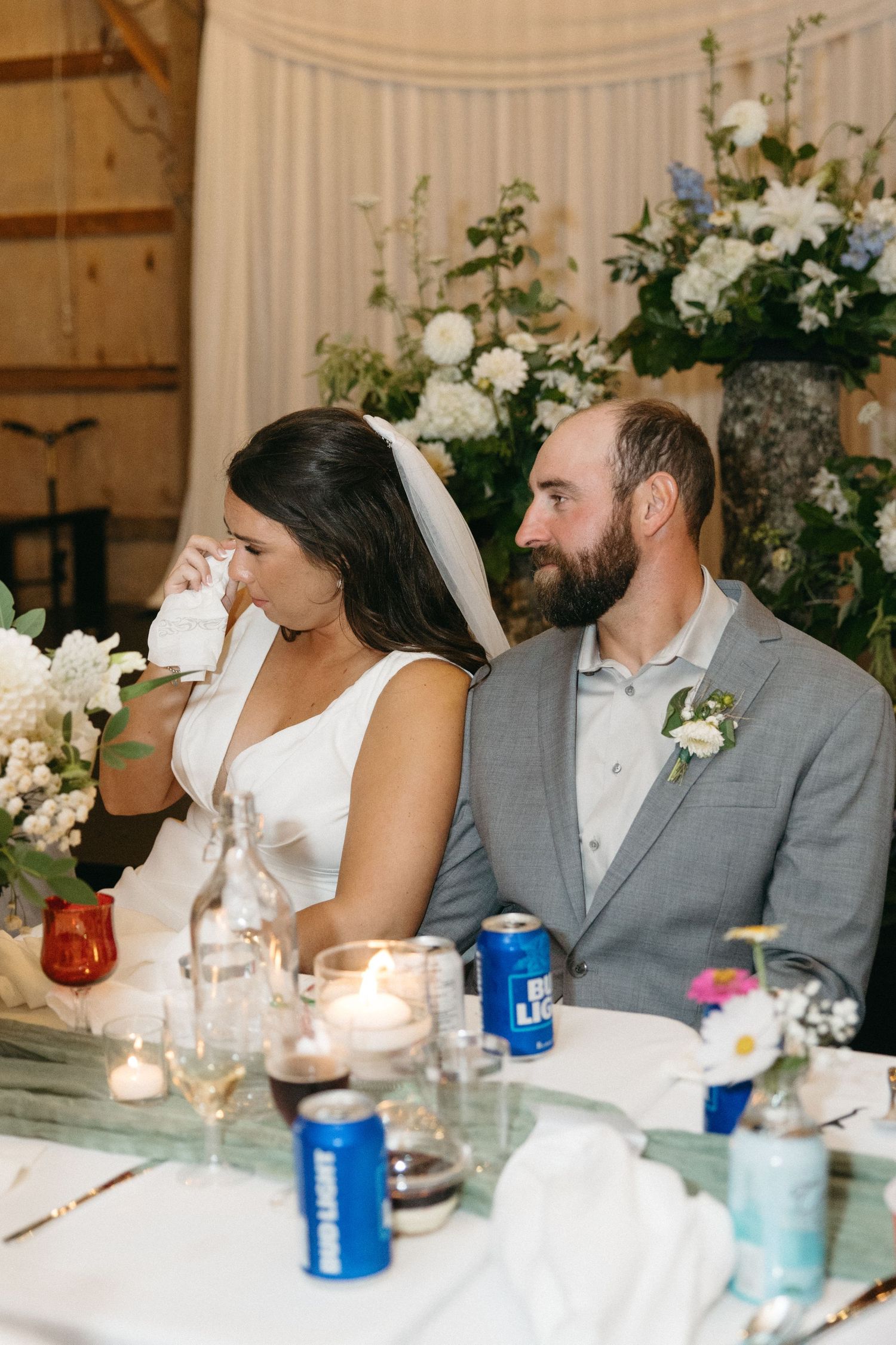 Wedding couple sits at reception table decorated with white floral arrangements and candles in rustic barn venue.