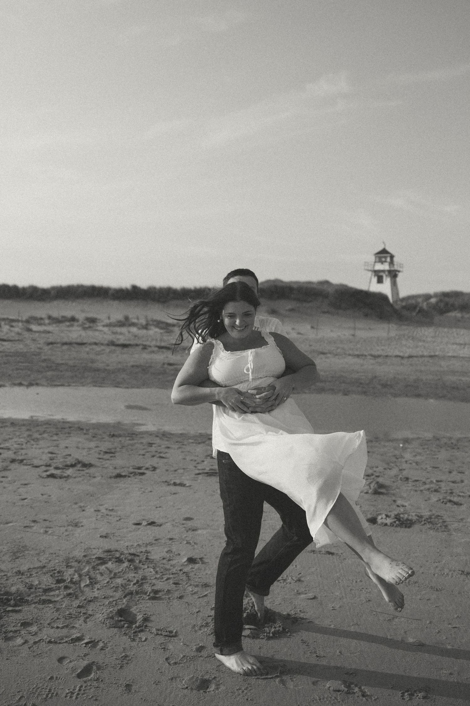 A playful black and white photo of a couple dancing on the beach with a lighthouse in view.