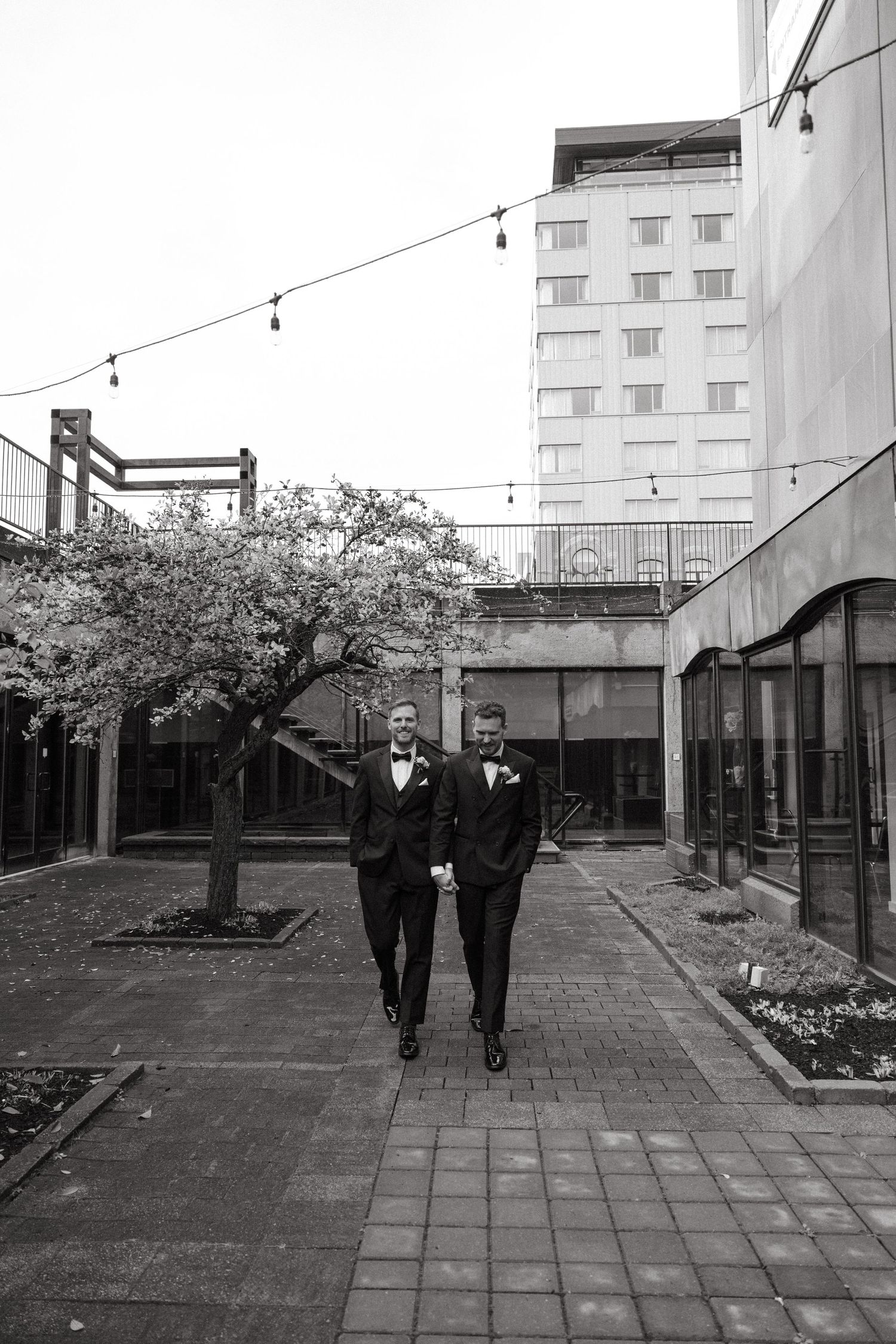Two people in formal attire walk together through an outdoor courtyard with trees and buildings.