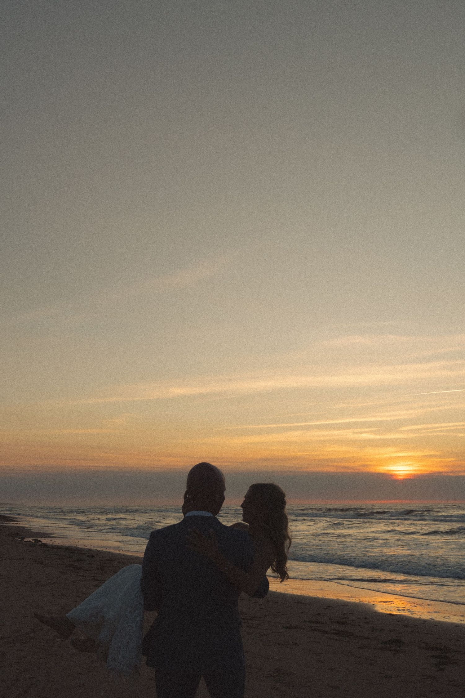 Two people watching a vibrant orange sunset on a beach with waves rolling in.