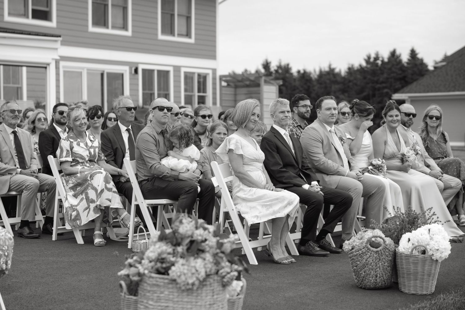 Black and white photo of wedding guests seated in white chairs at an outdoor ceremony.